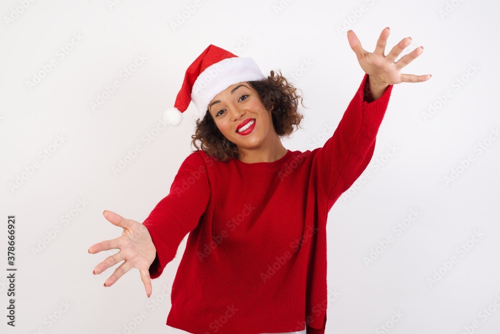 Young arab woman with curly hair wearing christmas hat on white background looking at the camera smiling with open arms for hug. Cheerful expression embracing happiness.