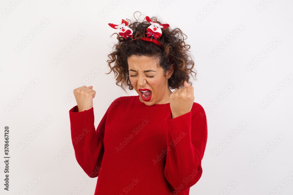 Young arab woman with curly hair wearing christmas headband standing on white back very happy and excited doing winner gesture with arms raised, smiling and screaming for success. Celebration concept. Young arab woman with curly hair wearing christmas headband standing on white back very happy and excited doing winner gesture with arms raised, smiling and screaming for success. Celebration concept.