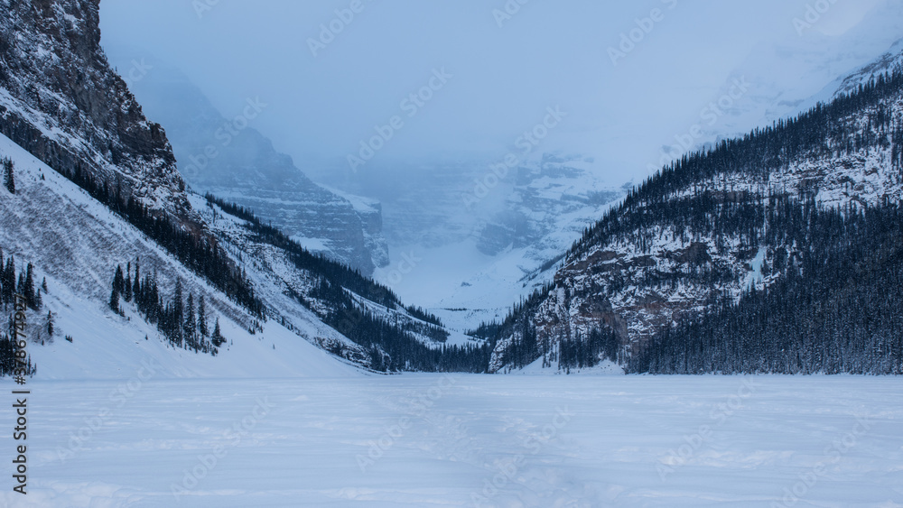 Obraz premium Snowy path on a frozen lake Canadian rocky mountain landscape and glacier mountain peak in the fog. Freezing cold and heavy snowfall at dusk. Majestic mountain range at Lake Louise, Alberta, Canada