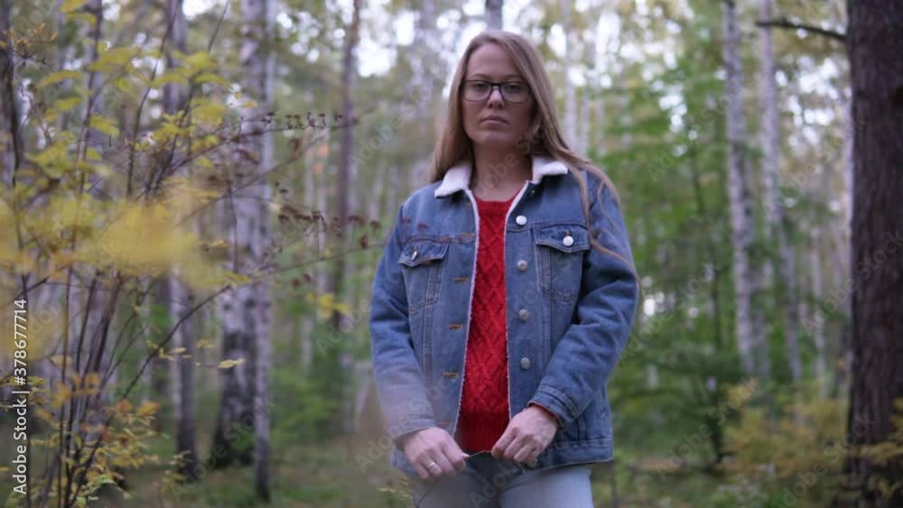 Pretty blonde girl in jeans jacket staying in an autumn birch forest. Slow motion rack focus medium shot panorama. Young woman behind yellow leaves bokeh, autumn evening. Pretty blonde girl in jeans jacket staying in an autumn birch forest. Slow motion rack focus medium shot panorama. Young woman behind yellow leaves bokeh, autumn evening.