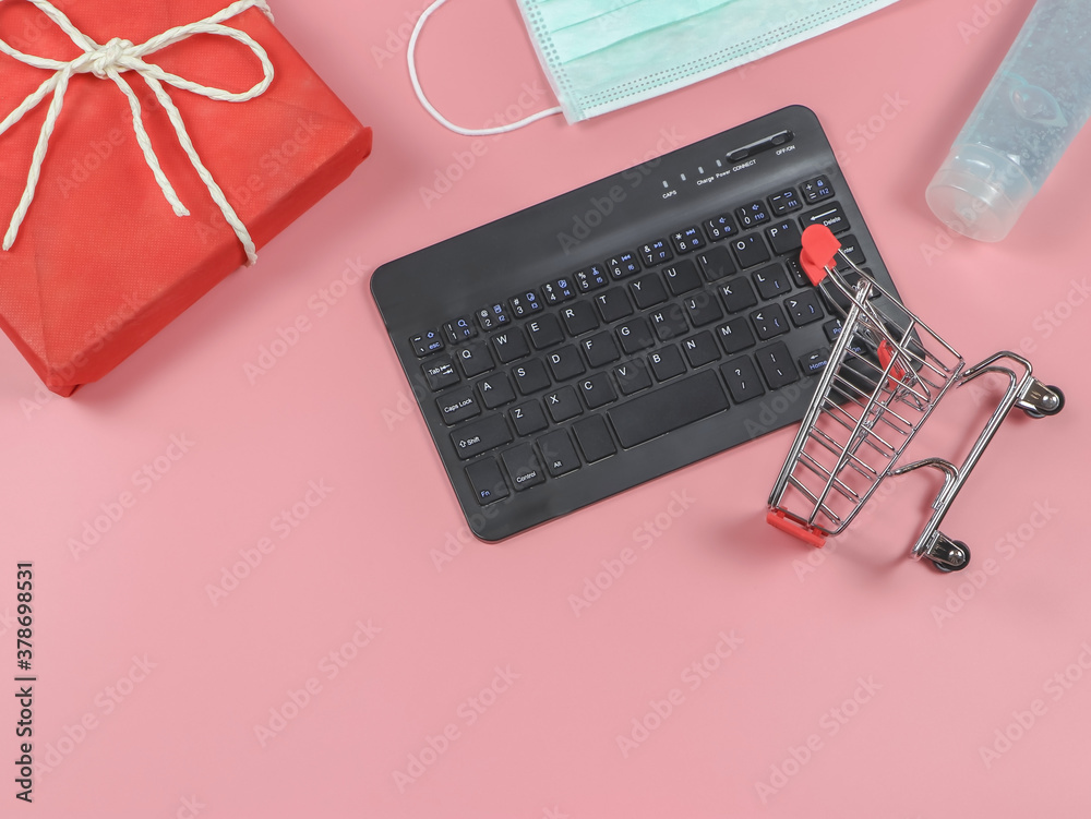 flat lay of computer keyboard , shopping cart,red gift box , medical mask  and alcohol sanitizer gel on pink background with copy space. Covid19  prevention, new normal and online shopping. Stock Photo