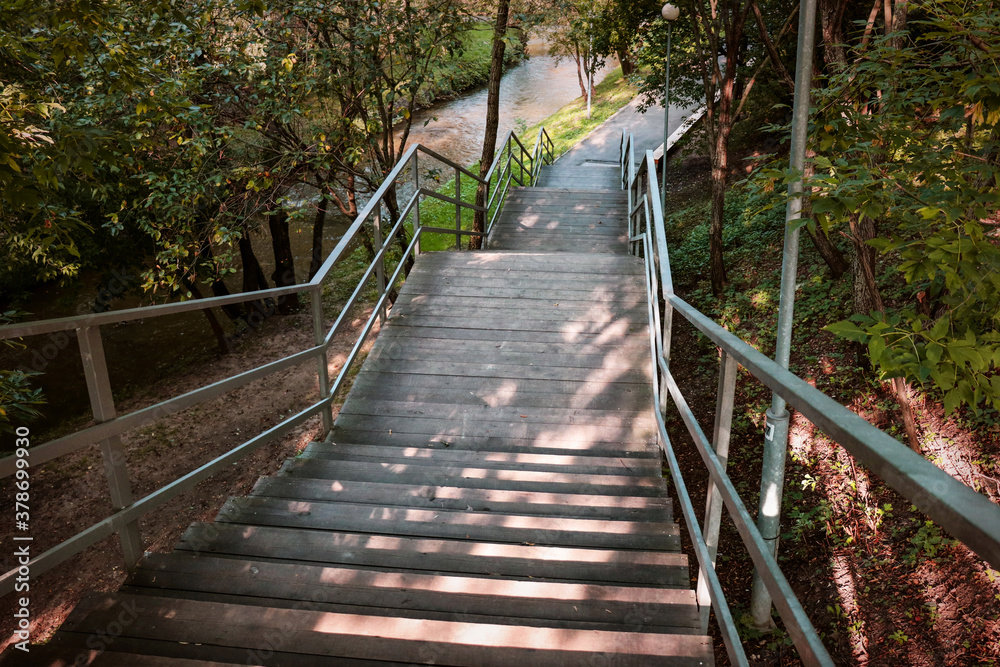 Ladder in park with metal handrails. Wooden steps going down among trees, river in background. Climbing stairs in fresh air, early autumn. Sunlight, shadows from foliage