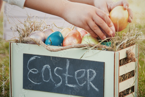 Unrecognizable child put colored eggs into wooden box full of multi colored painted easter eggs staying on the green grass. Easter holiday concept