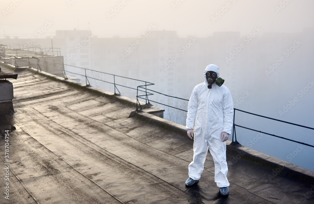Man wearing white coverall and gas mask, standing on the roof of an urban building with his back to a rising sun in foggy weather, concept of environmental problem