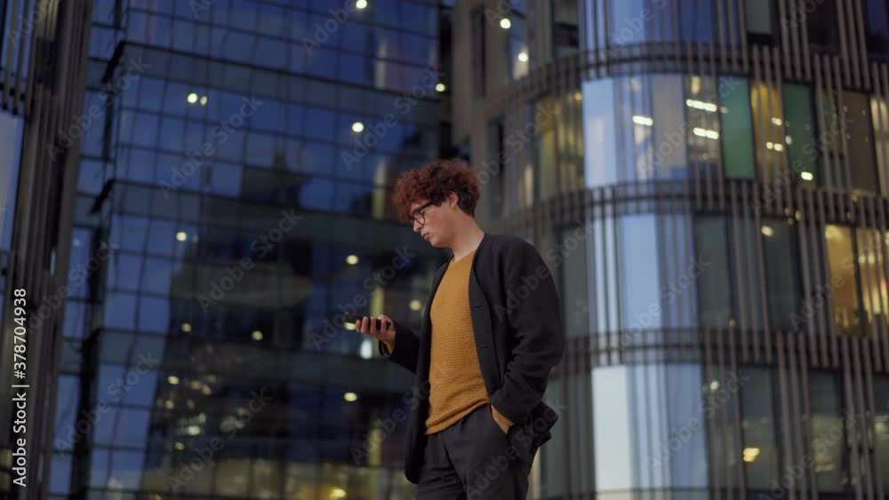 Side view medium shot of young man with curly red hair and in eyeglasses walking into frame, text messaging on cell phone and walking away against background of modern glass building Side view medium shot of young man with curly red hair and in eyeglasses walking into frame, text messaging on cell phone and walking away against background of modern glass building