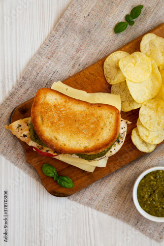 Homemade Pesto Chicken Sandwich with Potato Chips on a rustic wooden board on a white wooden surface, top view. Flat lay, from above, overhead.