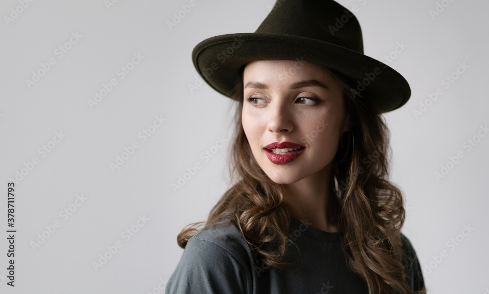 Close up portrait of female in stylish black hat with brown curls and wine red lips posing in studio isolated on gray background. Autumn fashion concept. Close up portrait of female in stylish black hat with brown curls and wine red lips posing in studio isolated on gray background. Autumn fashion concept.
