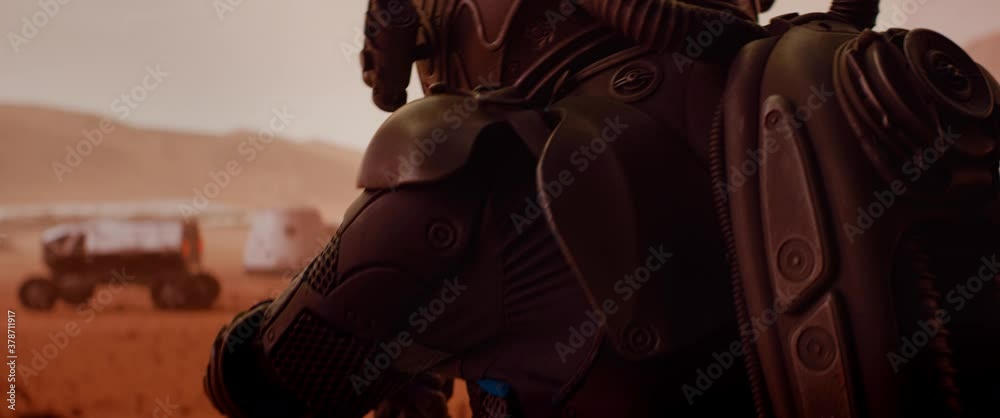 HANDHELD TRACKING back view of astronaut wearing space suit walking on a surface of a red planet. Martian base and rover in the background. Mars colonization concept