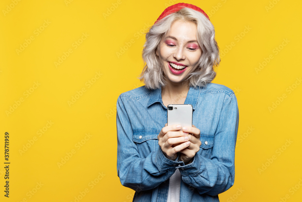 Portrait of smiling woman with modern smartphone in hands. Model wearing casual jeans coat and red head band. Copy space in left side. Isolated on yellow Portrait of smiling woman with modern smartphone in hands. Model wearing casual jeans coat and red head band. Copy space in left side. Isolated on yellow