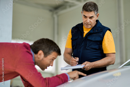 Auto mechanic taking notes while customer is showing his problem a car in repair shop.