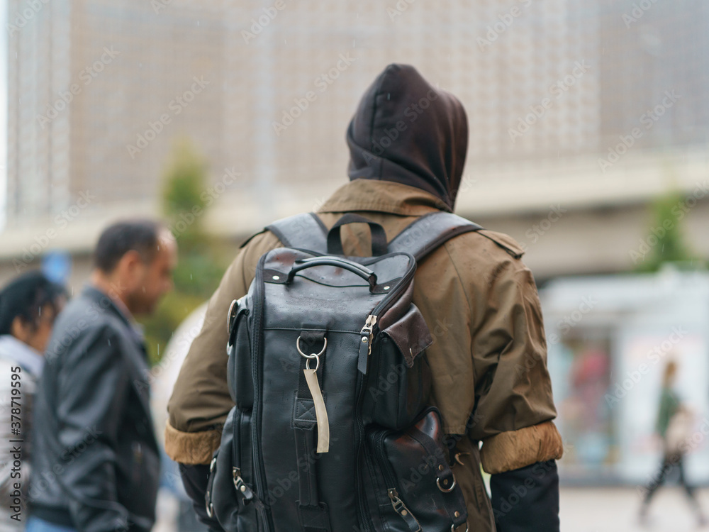Young man with black backpack at the autumn city street. Rainy day. He put on hoodie and rain coat. Modern fashion style of youth. Back / rear view. Coronavirus pandemic time. Young man with black backpack at the autumn city street. Rainy day. He put on hoodie and rain coat. Modern fashion style of youth. Back / rear view. Coronavirus pandemic time.