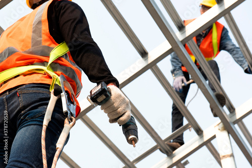 Construction workers wear safety straps while working on the building's roof structure at a construction site. Roofer using a pneumatic nail gun.