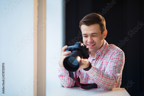 a male photographer in a checkered shirt sits at a table and holds a camera in his hands. creative profession or hobby. professional photo sessions.