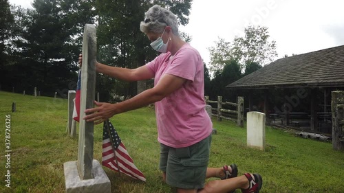 women wearing a mask kneels at gravestone after planting an american flag at site photo