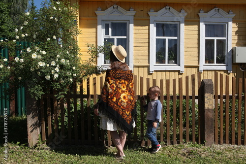 Russian girl in vintage Pavlovo Posad shawl and straw hat. Wooden rural house in Yukhnov town, Russia. Russian folk style in architecture. Architectural fashion. Summer countryside, blooming garden photo