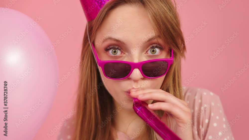 Girl Wearing A Pink Party Hat And Glasses Blowing A Party Blower While Holding A Pink Balloon. - close up shot Girl Wearing A Pink Party Hat And Glasses Blowing A Party Blower While Holding A Pink Balloon. - close up shot