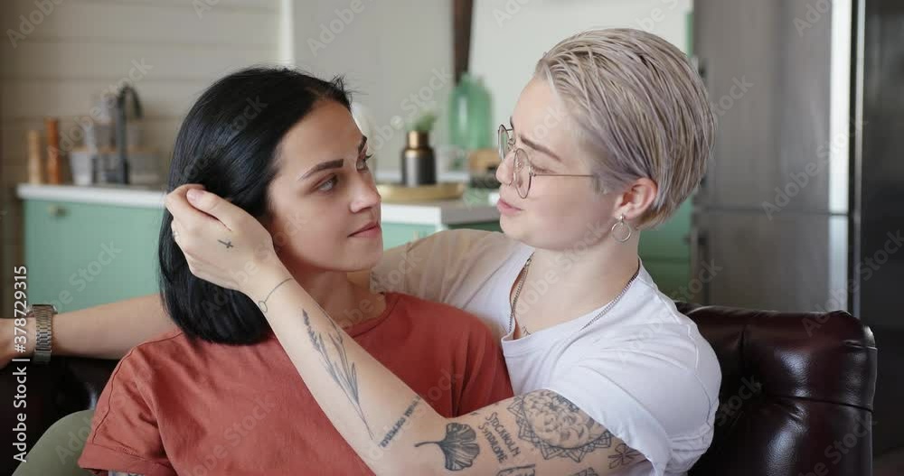 Young woman in glasses hugs smiling girlfriend with loose hair sitting on brown leather sofa at home slow motion close view Young woman in glasses hugs smiling girlfriend with loose hair sitting on brown leather sofa at home slow motion close view