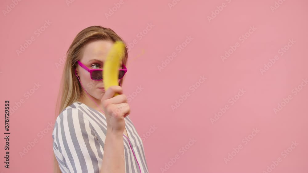 Pretty Woman Wearing Pink Framed Eyewear Moving Head Side To Side While Holding A Fresh Yellow Banana Like A Gun Against Pink Background - Medium Closeup Shot Pretty Woman Wearing Pink Framed Eyewear Moving Head Side To Side While Holding A Fresh Yellow Banana Like A Gun Against Pink Background - Medium Closeup Shot