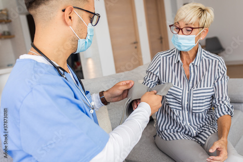 Male nurse measures blood pressure to senior woman with mask while being in a home visit.