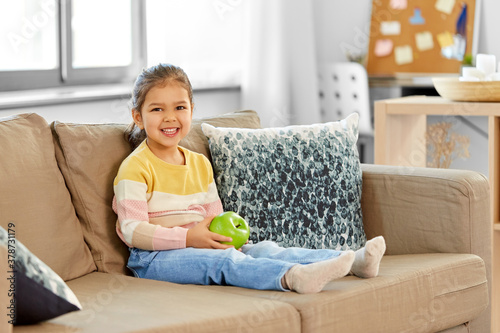 healthy eating and childhood concept - happy smiling little girl with green apple sitting on sofa at home