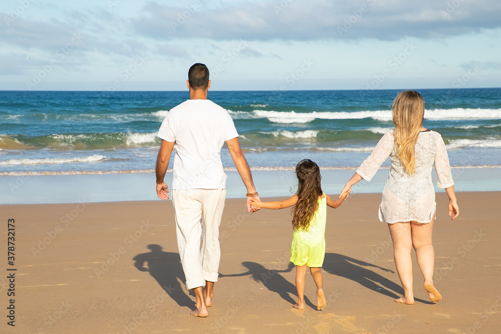 Young man, woman and kid in summer clothes walking on wet sand to sea, spending leisure time on beach. Rear view. Family outdoor activities concept
