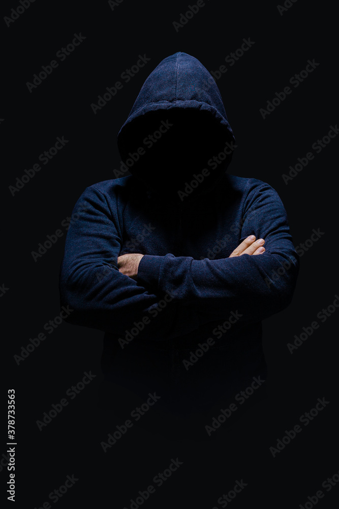 Man in dark blue hoodie, face hidden in shadow, unrecognizable, crossed arms, alone in dark 