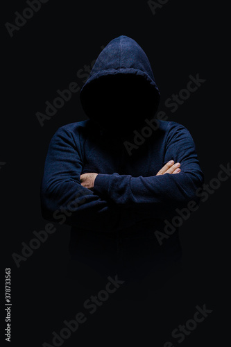 Man in dark blue hoodie, face hidden in shadow, unrecognizable, crossed arms, alone in dark 