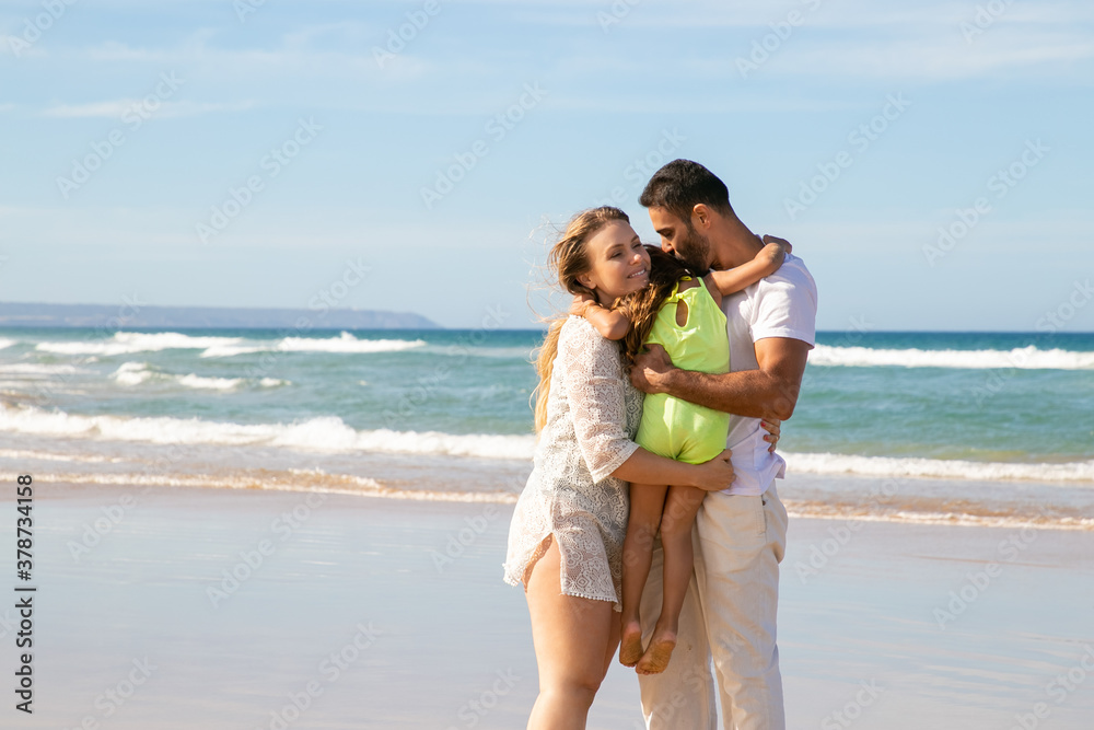 Happy young couple of parents holding little daughter in arms, hugging and kissing girl. Mom, dad and kid enjoying leisure time on beach at sea. Parenting and childhood concept Happy young couple of parents holding little daughter in arms, hugging and kissing girl. Mom, dad and kid enjoying leisure time on beach at sea. Parenting and childhood concept