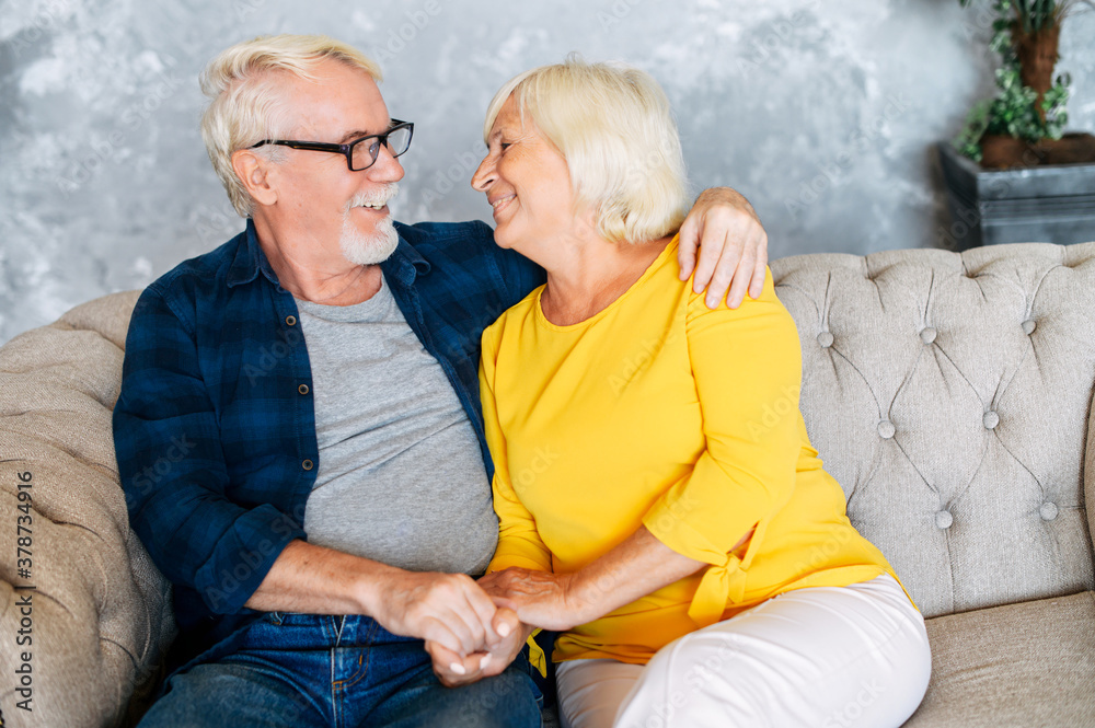 Elderly couple looks to each other eyes with love and care spending leisure at home. A bearded senior husband in eyeglasses and gray-haired beautiful wife holding hands sitting on the sofa Elderly couple looks to each other eyes with love and care spending leisure at home. A bearded senior husband in eyeglasses and gray-haired beautiful wife holding hands sitting on the sofa