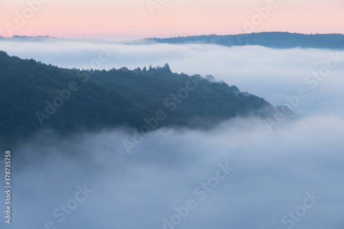 Mountains in fog at beautiful morning in autumn. Landscape with mountain valley, low clouds, forest, colorful sky , nature illuminated at dusk. Dramatic mist in forest at sunrise, space for text