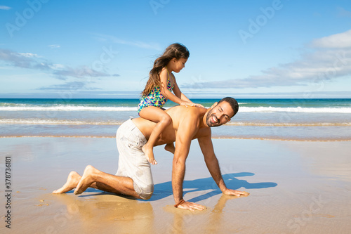 Cheerful dad going on hands and knees on beach, carrying little girl on his back. Side view. Family outdoor activities concept
