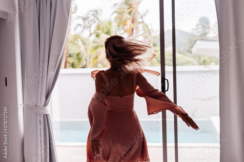 Happy caucasian calm stylish cute woman in pink summer dress and elegant brown headband standing on teracce at villa enjoying beautiful day alone.   © Анастасия Каргаполов