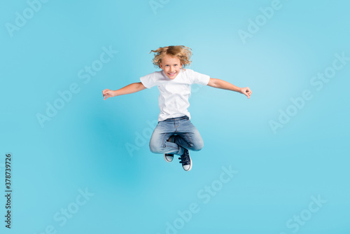 Photo of small child jump funny look camera raise hands wear white shirt jeans sneakers isolated blue color background