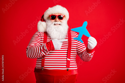 Photo of old man grey beard hold direct finger paper plane smile wear santa claus x-mas costume suspenders sunglass striped shirt cap isolated red color background © deagreez