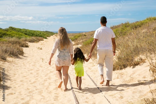Parents and kid wearing summer clothes, walking along sand path to sea, girl holding parents hands. Back view. Family outdoors concept © Mangostar