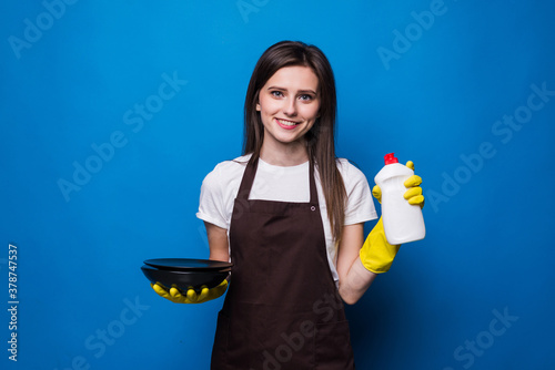 Canvastavla Young woman in orange apron with washed plate showing sponge and dishwashing detergent isolated on blue background
