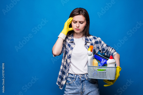 Fotografie Young housewife in apron with a basket with cleansers and brushes feel tired on blue background