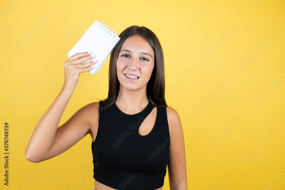 Beautiful young girl kid over isolated yellow background smiling and showing blank notebook in her hand Beautiful young girl kid over isolated yellow background smiling and showing blank notebook in her hand