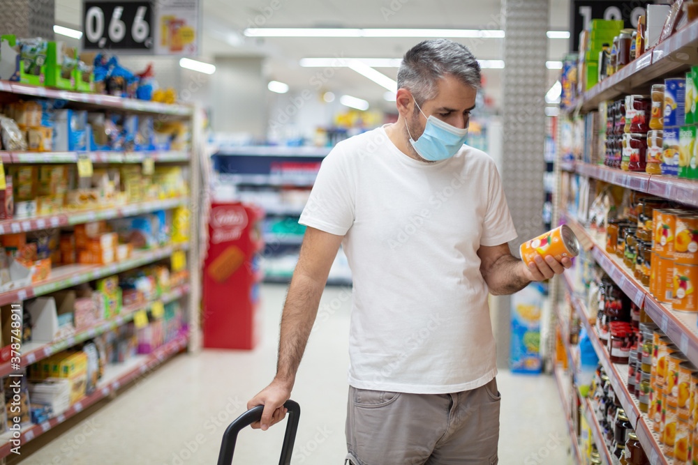 A man in a medical mask in a supermarket holds canned food in his hand.