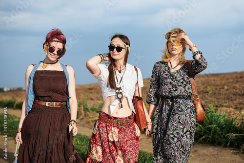 Three hippie women, wearing boho style clothes, walking on dirt road on green field, having fun. Female friends, traveling together in countryside. Eco tourism concept. Summer leisure free time.