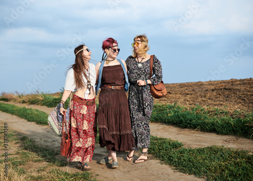 Three hippie women, wearing boho style clothes, walking on dirt road on green field, having fun. Female friends, traveling together in countryside. Eco tourism concept. Summer leisure free time.