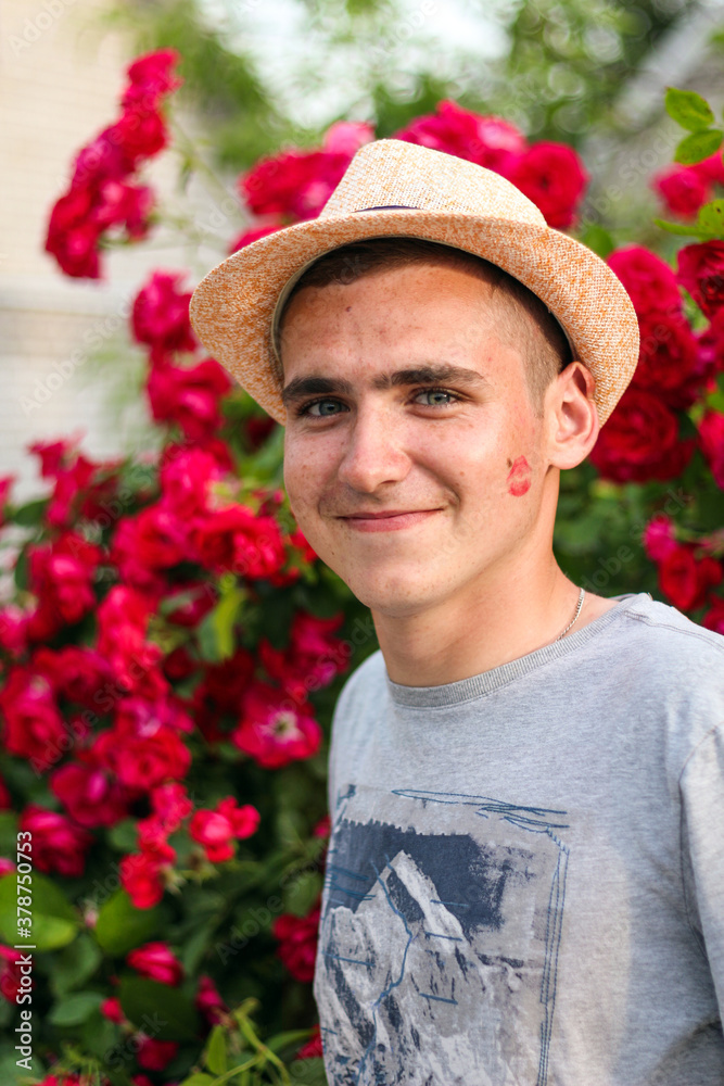 Portrait of handsome brunet guy smiling excited, express own romantic feelings or sympathy. Charming young guy with a print of women's lips on his cheek standing by flowers in Garden. Selective focus.