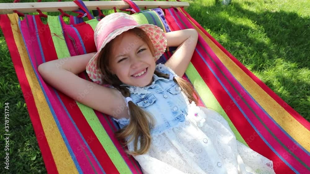 a little girl lying in a hammock on the street in the park. a little sweet girl in a hat lying on a bright hammock. girl in the park, garden