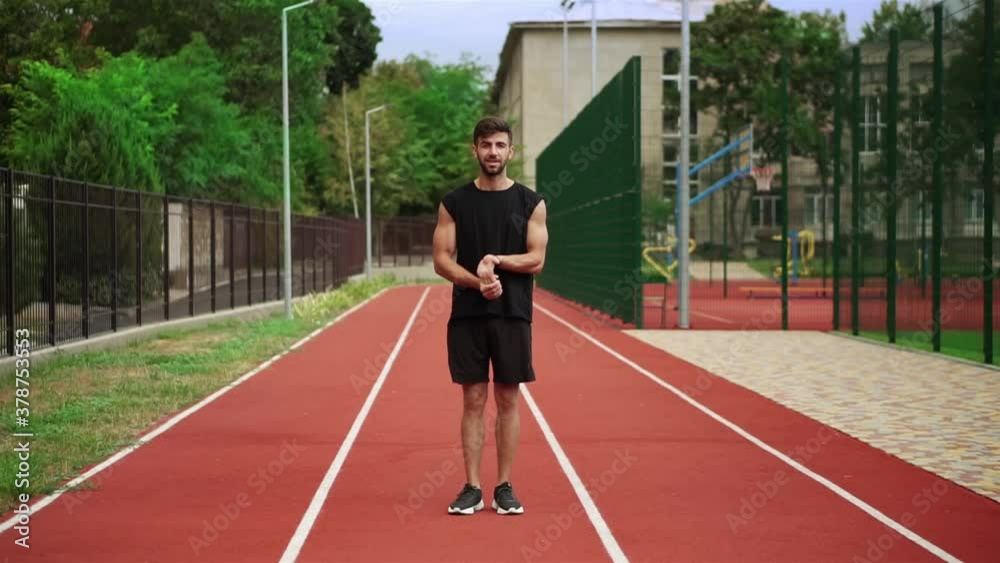 Fit, bearded man standing on running track outdoors on modern stadium warming up his wrists Fit, bearded man standing on running track outdoors on modern stadium warming up his wrists