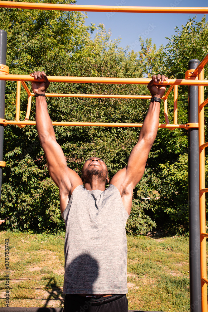 Handsome fit African American young man doing pull-ups on chin-up bar during workout outdoors Handsome fit African American young man doing pull-ups on chin-up bar during workout outdoors