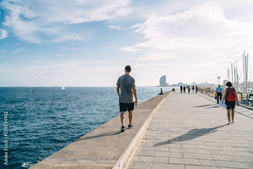 Back view of caucasian male runner recreating walking on quay near sea water in city harbor on sunny morning, sportsman having break after cardio workout training outdoors for keeping body shape