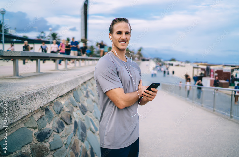 Half length portrait of smiling muscular male jogger enjoying healthy lifestyle listening audiobook via smartphone and earphones, handsome man runner listening playlist mucin on mobile phone outdoors