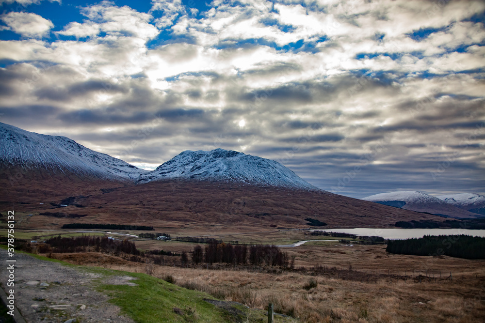 Scottish Highlands in fall. Rural view with river, lake, forest, house and mountains. Peaks with snow. Blue sky with clouds.