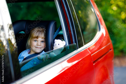 Bilde på lerret Adorable toddler girl sitting in car seat, holding plush soft toy and looking out of the window on nature and traffic