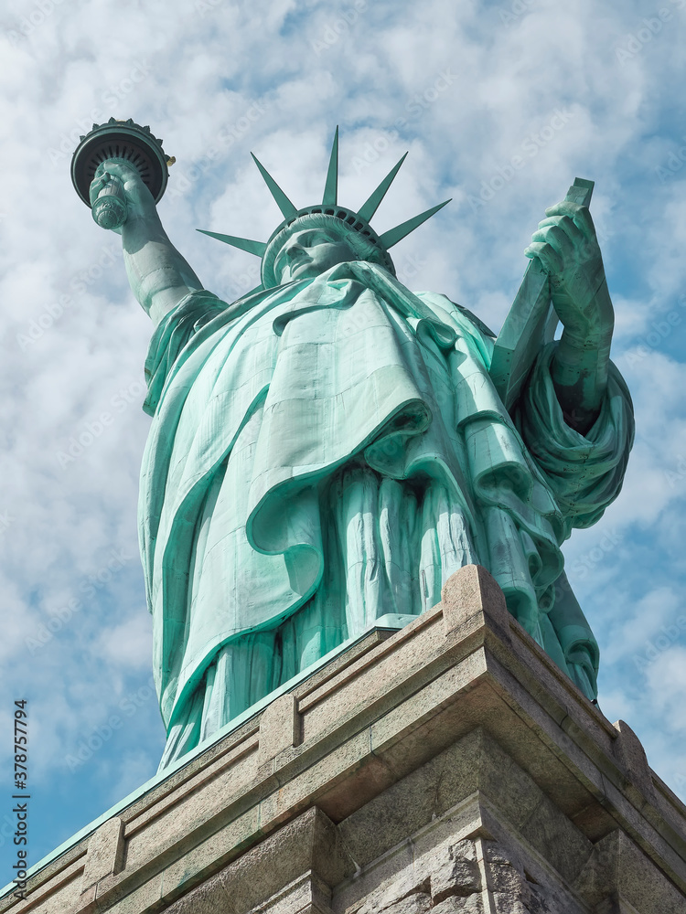 Fototapeta premium vista en contrapicado de la Estatua de la libertad y parte de su pedestal, con el fondo de cielo azul turquesa con nubes dramaticas, en Nueva York, Estados Unidos
